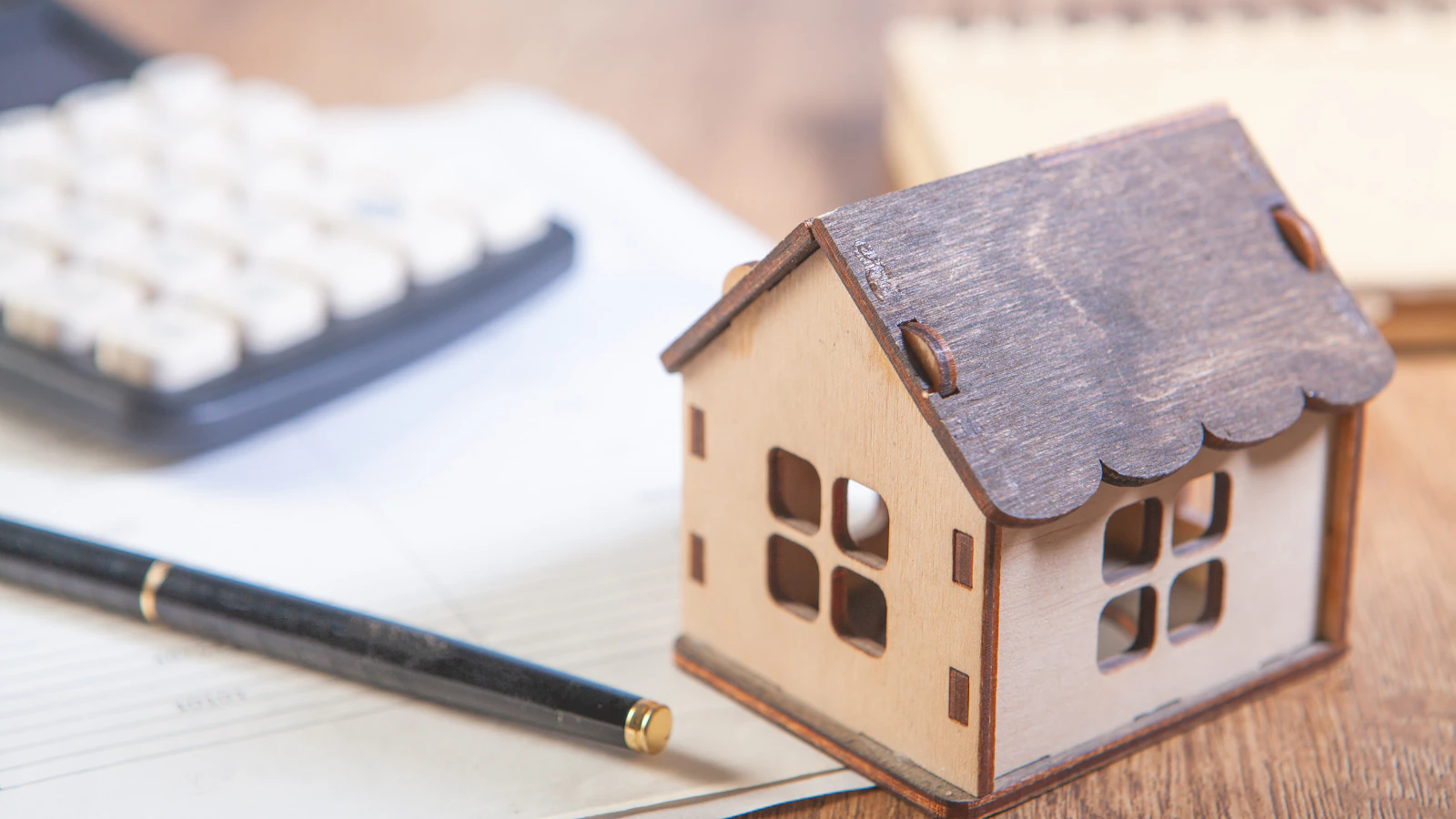 Wooden model home beside a calculator, notebook and pen on a desk.